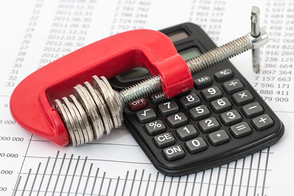 Wood clamp secures coins, calculator nearby, illustrating savings with monoline workers' comp.